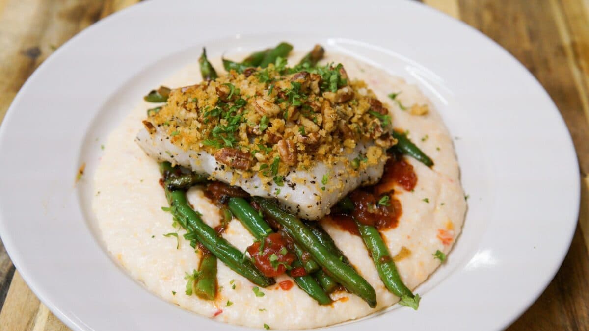 Plated dish featuring a pecan-crusted white fish fillet served over sautéed green beans, tomato chutney, and creamy cheese grits, garnished with fresh herbs.